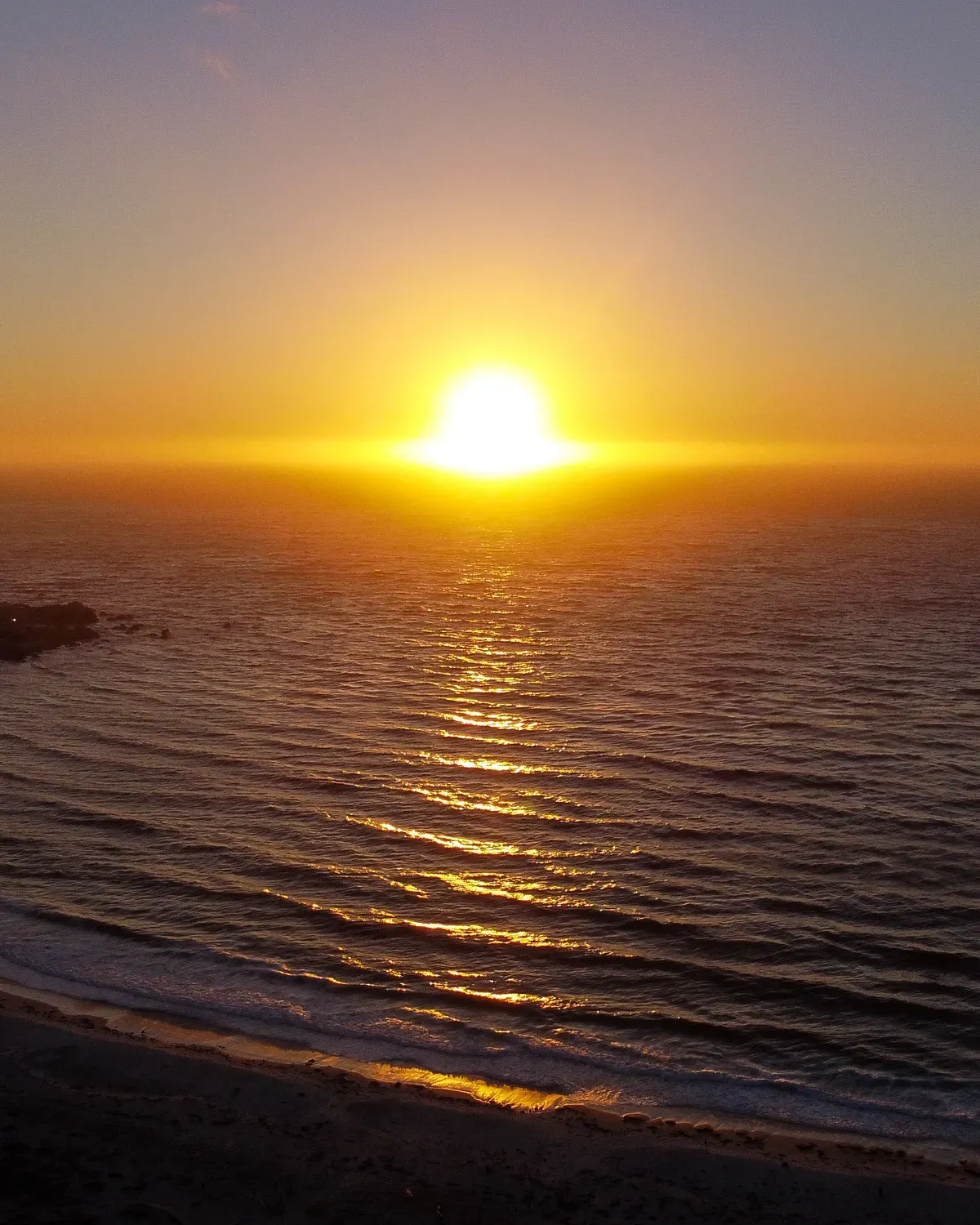 A long beach seen from the air, parallel lines of swell arriving on the sand in the morning haze
